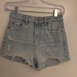 Ljusa jeansshorts från Zara - Snygga ljusblå jeansshorts från Zara med hög midja och slitna detaljer. Rå kant nertill och flera coola revor framtill och baktill ger en trendig look. Klassisk femficksmodell i mjukt denimtyg, perfekta för sommardagar.