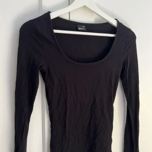 Svart långärmad topp Gina Tricot - En svart långärmad topp från Gina Tricot med rundad, djup urringning. Tunn och mjuk i bomullsmix, perfekt att styla med jeans eller kjol. Enkel och stilren design som funkar till många looks.