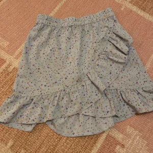 Ljusblå blommig kjol med volang från PIECES - Supersöt ljusblå kjol från PIECES med små blommor i olika färger. Kjolen har omlott-look och volangdetaljer längs nederkanten och framtill. Resår i midjan för extra komfort. Perfekt för dig som gillar en feminin stil.