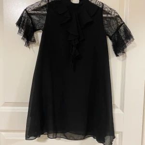 Svart klänning med spetsärmar och volang - Svart klänning med korta spetsärmar och volangdetaljer framtill. Klänningen har en lös passform och är tillverkad i ett lätt, flödande material. Perfekt för dig som gillar romantiska detaljer och en elegant look.