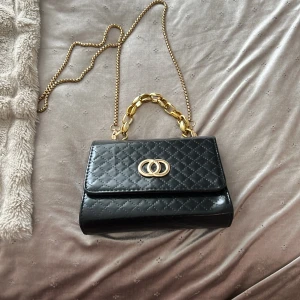 Svart quiltad axelväska med guldkedja - Säljer en svart quiltad axelväska med guldfärgade detaljer och kedja. Väskan har ett elegant mönster, snyggt spänne framtill och innerfack med dragkedja. Perfekt accessoar för att lyfta din outfit.