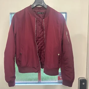 Vinröd bomberjacka från stradivarius  - Säljer en vinröd bomberjacka från stradivarius  med quiltat innerfoder. Jackan har dragkedja framtill, fickor med tryckknappar och en ärmficka med dragkedja. Klassisk bombermodell med ribbade muddar vid ärmslut och midja.