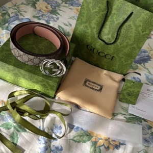 Gucci GG Supreme bälte med silverfärgat spänne - Säljer ett Gucci GG Supreme bälte med klassiskt monogrammönster och stort silverfärgat GG-spänne. Bältet har beige och bruna toner med insida i ljusrosa. Kommer med originalförpackning, dustbag och kvitto.
