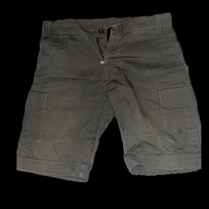 Gröna coola cargoshorts från Adidas - Säljer ett par gröna cargoshorts från Adidas med klassiska cargofickor på sidorna och broderad logga. Shortsen har normal passform och stängs med knapp och dragkedja. Perfekta för en cool stilrent stil ! 
