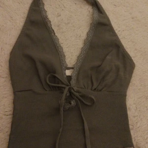 Halterneck-linnetopp  - Säljer en mörk grön/oliv färgad ribbad halterneck-linnetopp från Pull & Bear. Toppen har spetskant längs urringningen och knytning framtill. Storlek XS, bra skick💗