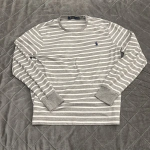 Gråvit randig långärmad tröja från Polo Ralph Lauren - Säljer en stilren grå och vit randig långärmad tröja från Polo Ralph Lauren. Tröjan har rund halsringning och den klassiska broderade loggan på bröstet. Perfekt för en avslappnad och snygg look.