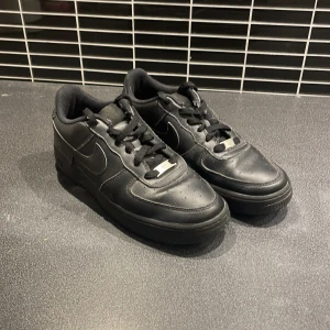 Svarta Nike Air Force 1 sneakers - Säljer ett par klassiska helsvarta Nike Air Force 1 sneakers med snörning och rund tå. Skorna har en platt sula och är tillverkade i skinn. Perfekta för dig som gillar en clean still.