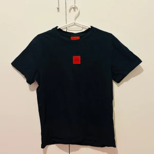 Svart t-shirt från Hugo Boss - Säljer en svart t-shirt från Hugo Boss med röd Hugo-logga på bröstet. Klassisk passform och rund halsringning. Perfekt för dig som gillar stilrena plagg med en touch av designer.