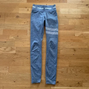 Grå träningstights från Stronger - Snygga grå träningstights från Stronger med hög midja och vit randdetalj på ena benet. Tightsen är stretchiga och har en bekväm passform, perfekta för gymmet eller löpning. 