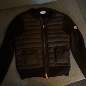 Svart cardigan från Moncler - Säljer en svart cardigan från Moncler med quiltade paneler framtill, två fickor med lock och dragkedja. Jackan har långa ärmar i stickat material och klassisk Moncler-logga på ärmen. Perfekt för kyliga dagar.