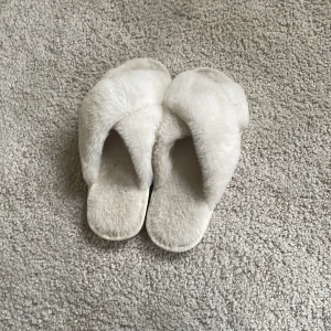 Vita fluffiga tofflor - Mysiga vita tofflor med korsad design i mjukt, fluffigt material. Perfekta att ha hemma för extra komfort och värme. Säljes i storlek 38-39.