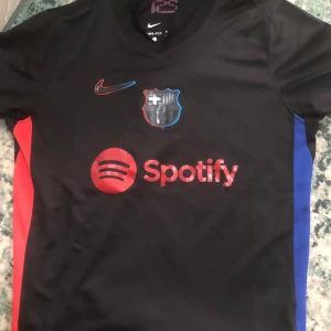  FC Barcelonas tröja  - Säljer en svart FC Barcelona fotbollströja från Nike med Spotify-logga, klubbmärke på bröstet.. Tröjan har röda och blå detaljer på sidorna samt UNHCR-tryck på ryggen. Priset kan diskuteras pm