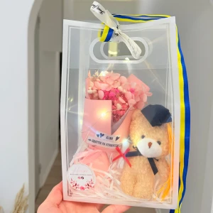 Studentpresent med nalle och rosa blommor - Söt presentpåse med en liten brun nallebjörn med blå studentmössa och en bukett rosa blommor i pappersförpackning. Perfekt som gåva till studenten. Påsen har blågult band och dekorativ rosett.