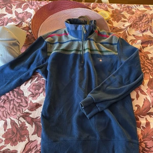 Blå halvzip tröja från GANT - Säljer en blå långärmad tröja från GANT med halv dragkedja och ståkrage. Tröjan har gråa, gröna, blå och röda ränder över bröstet och axlarna. Perfekt för en avslappnad och stilren look.