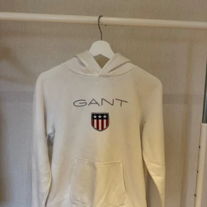Vit hoodie från GANT med logga - Säljer en vit hoodie från GANT med broderad logga och sköld på bröstet. Tröjan har huva och en stor magficka framtill. Klassisk och stilren design med mjukt material, perfekt för dig som gillar bekväma och snygga plagg.