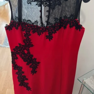 Röd cocktail klänning med svart spets - Säljer en elegant röd cocktail klänning med svart spetsdetalj upptill och längs ena sidayn. Klänningen har transparent mesh upptill med blommig spets och dekorativa knappar i ryggen. Perfekt för speciella tillfällen.
