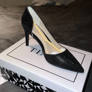 Svarta glittriga klackar från tiamo - Säljer ett par eleganta svarta pumps från Tiamo med glittrigt material och spetsig tå. Skorna har hög klack och en klassisk siluett som passar perfekt till festliga tillfällen.
