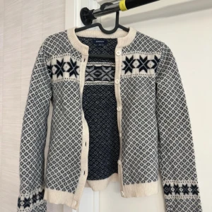 Mönstrad ullkofta från Gant - Säljer en klassisk stickad kofta från Gant i blått och vitt med traditionellt mönster och dekorativa knappar vid ärmsluten. Koftan har rund halsringning, långa ärmar och knäppning framtill. 100% lammull. Använd en gång