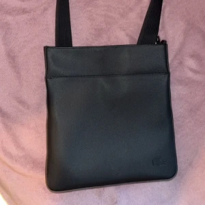 Svart axelväska från Lacoste - Säljer en stilren svart axelväska från Lacoste med justerbart axelband. Väskan har minimalistisk design och diskret Lacoste-logga i nedre hörnet. Perfekt för att bära det viktigaste i vardagen.