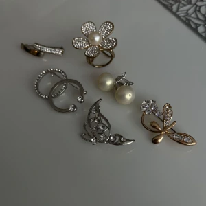 Smycken - Säljer ett set med olika smycken: ett blommigt ring med pärla, ett par pärlörhängen, en guldfärgad fjärilsbrosch med strass, två ringar med stenar och ett silvrigt brosch med strass. Perfekt för dig som gillar unika accessoarer. hör av er vid intresse, priset är allting tillsammans💞