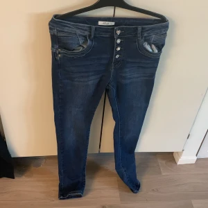 Mörkblå jeans med dekorativa knappar  - Mörkblå jeans från Jewelly. Byxorna har fyra dekorativa knappar framtill, varav en är hjärtformad och de andra har pärl- och strassdetaljer. Klassiska bakfickor och lätt tvättad look.