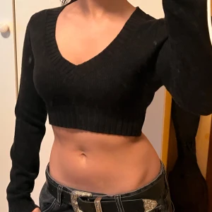 Svart croppad stickad tröja från H&M Divided - Säljer en svart croppad stickad tröja från H&M Divided. Tröjan har v-ringning och långa ärmar med ribbade muddar. Perfekt för dig som gillar en trendig och enkel stil.