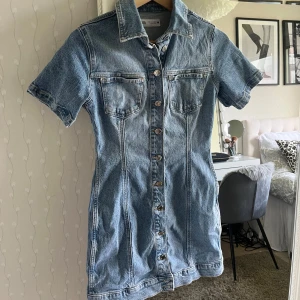 Kort jeansklänning med knappar - Säljer en snygg kort jeansklänning med krage och korta ärmar. Klänningen har knappar hela vägen framtill och två bröstfickor. Figurnära passform som framhäver siluetten. Oanvänd och väldigt bra skick!🥰