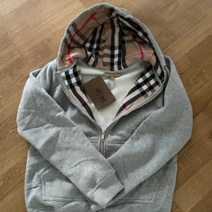 Grå Burberry zip med rutigt foder - Säljer en grå hoodie från Burberry med dragkedja och klassiskt rutigt foder i huvan. Tröjan har broderad logga på bröstet och ribbade muddar. Perfekt för dig som gillar stilrena och exklusiva plagg.