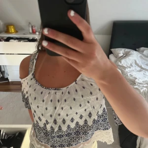 Vit och blå croppad blus med spetsdetaljer - Säljer en söt croppad blus i vitt med blått mönster och tunna axelband. Blusen har fina spetsdetaljer och volang längst ner, perfekt för varma dagar.