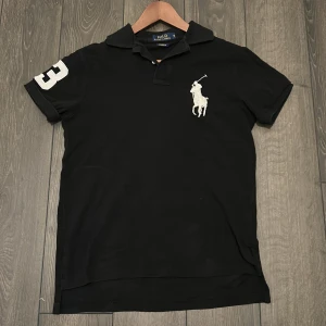 Svart pikétröja från Polo Ralph Lauren - Svart pikétröja från Polo Ralph Lauren med klassisk krage, broderad vit polospelare på bröstet och stor vit siffra 3 på ena ärmen. Custom fit-modell som passar till många tillfällen. Du kommer inte att bli besviken! Skriv om du har några frågor 👏!