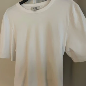 Vit croppad t-shirt med rosett från COS - Vit t-shirt från COS i bomull med korta ärmar och rund halsringning. Baktill finns en stor rosett och modellen är croppad vilket ger en trendig look. Perfekt för sommaren och enkel att styla till olika outfits.