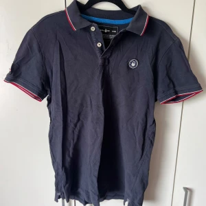 Mörkblå pikétröja med röd och vit kant - Säljer en mörkblå pikétröja från Sail racing med korta ärmar och klassisk krage. Tröjan har röda och vita detaljer på krage och ärmslut samt en broderad logga på bröstet. 