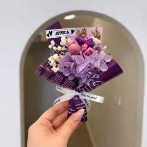 Jessica bukett  - En liten, vackert bunden bukett med torkade blommor i lila och rosa toner, inslagen i papper med texten 'Jessica' på en etikett. Perfekt som dekoration eller personlig present.