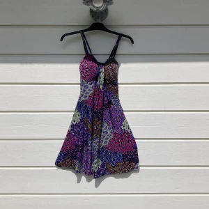 Färgglad kortklänning med mönster - Säljer en färgglad kortklänning med smala axelband och ett livfullt, abstrakt mönster i lila, rosa, blått och grönt. Klänningen har en lätt utställd form och är perfekt för varma dagar.