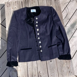 Svart kavaj med sammetsdetaljer från Perry Landhaus - Stilren mörk kavaj med sammetskrage och manschetter från Perry Landhaus. Kavajen har dekorativa silverfärgade knappar framtill och två fickor. Klassisk passform och elegant design, perfekt för dig som gillar en tidlös look.