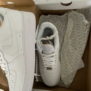 Vita Nike Air Force 1 sneakers - Klassiska helvita Nike Air Force 1 sneakers med rund tå och platt sula. Skorna har snörning och är tillverkade i syntetmaterial med diskreta detaljer. Perfekta för en clean och stilren look.