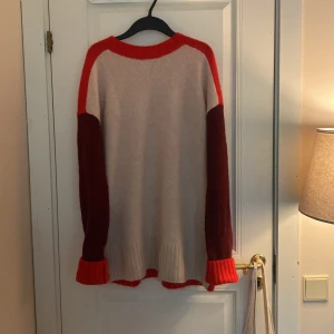 Röd och beige stickad tröja - Säljer en stickad tröja i rött och beige med mörkröda ärmar och röda detaljer vid hals och muddar. Tröjan har lång ärm och är tillverkad i en mjuk ullblandning. Perfekt för kyliga dagar. Tröjan är lång så man kan ha den över ett par tajts eller jeans men funkar också som klänning om man är kort.