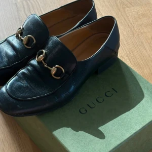 Svarta loafers från Gucci med gulddetaljer - Klassiska svarta loafers från Gucci i skinn med ikoniska guldfärgade hästbitsdetaljer framtill. Skorna har rund tå och låg klack, samt en elegant och tidlös design. Perfekta för dig som gillar lyxiga och stilrena skor.                        Serienummer: 571050 37 042