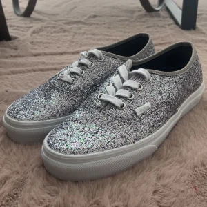 Glittriga sneakers från Vans - Säljer ett par silvriga glittriga sneakers från Vans med vita snören och vit sula. Skorna har rund tå och klassisk Vans-logga på sidan. Perfekta för dig som vill sticka ut med lite extra glitter i vardagen.Dem är aldrig använda.