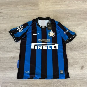 Inter Milan Sneijder 10 Champions League Finaltröja - Inter Milan matchtröja med Sneijder 10 på ryggen, blå och svart med vita detaljer. Champions League-märke på ärmen, Pirelli sponsortryck och klubbmärke fram. Final Madrid 2010 broderat på bröstet. Klassisk randig design och kortärmad modell.