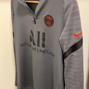 PSG Tröja - Säljer en grå långärmad träningströja från Nike med PSG-logga då jag inte använder längre. Perfekt för fotbollsträning eller som en snygg vardagströja. Storlek M men liten i storlek så kan även passa för dem med S.