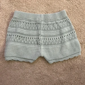Ljusblå stickade shorts - Säljer ett par ljusblå stickade shorts med virkat mönster och elastisk midja. Shortsen har en söt, vågig kant nertill och är perfekta för en avslappnad och trendig look. Tyvärr har de ett hål baktill (går säkert att laga) och en fläck framtill men syns ej så mycket (se bilder)💕