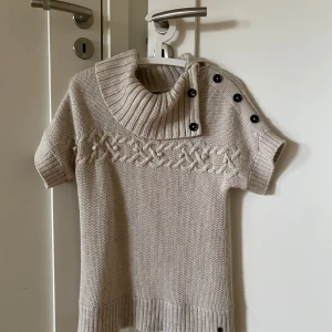 Beige stickad kortärmad tröja från s.Oliver - Beige stickad tröja från s.Oliver med korta ärmar, hög krage och dekorativa knappar på axeln. Snyggt flätmönster framtill och ribbade muddar vid ärmslut och nederkant. Perfekt för lager-på-lager och en stilren look! Jättemysig och assnygg!!❤️‍🔥❤️‍🔥kom med frågor!