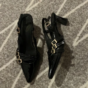 Svarta lackade klackskor med spännen - Säljer ett par svarta lackade klackskor med spetsig tå och tre dekorativa guldfärgade spännen på varje sko. Skorna har öppen häl och en elegant klack, perfekta för att lyfta din outfit.