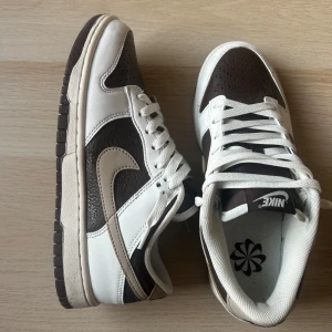 Nike Dunk Low sneakers i brunt och vitt - Säljer ett par Nike Dunk Low sneakers i brunt och vitt med beige swoosh. Skorna har klassisk låg modell, snörning och platt sula. Perfekta för dig som gillar streetwear och vill ha en stilren sneaker med snygga kontraster.
