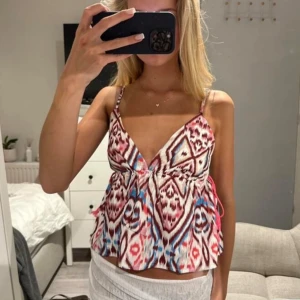 Mönstrad  linnetopp med tofsar - Säljer en färgglad croppad linnetopp med smala axelband och v-ringning. Toppen har ett iögonfallande mönster i rosa, blått och vitt samt dekorativa tofsar på sidan. Perfekt för sommardagar!
