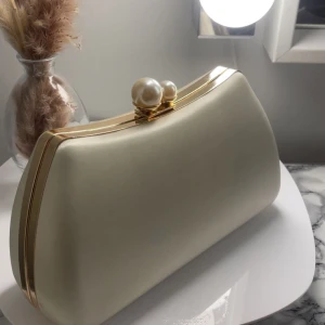 Vit kuvertväska med pärldetalj - Elegant vit kuvertväska med guldfärgade detaljer och ett unikt lås prytt med två pärlor. Väskan har en stilren, slät yta och en klassisk form som passar perfekt till speciella tillfällen. använd en gång men finns några små skavanker på