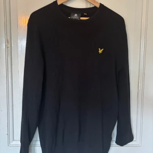 Lyle & Scott tröja - Klassisk och trendig svart tröja från Lyle & Scott med rund halsringning och den ikoniska gula loggan broderad på bröstet. Tröjan har lång ärm och är tillverkad i mjukt material som passar perfekt till många tillfällen.