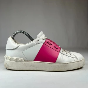 Valentino Open sneakers med rosa detalj - Säljer ett par vita Valentino Open sneakers med en bred rosa rem över mitten och nitar på hälen. Skorna har snörning och är tillverkade i skinn. Perfekta för dig som vill ha en stilren men ändå färgstark look.