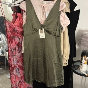 Olivgrön omlottklänning från Bik Bok - Säljer en olivgrön klänning från Bik Bok i omlottmodell med v-ringning. Klänningen har ärmlös design och är tillverkad i ett glansigt, mjukt material som ger en elegant känsla. Perfekt för dig som gillar stilrena och trendiga plagg.